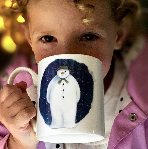 White mug with Raymond Briggs’ The Snowman character printed on the side. Mug held by a young person