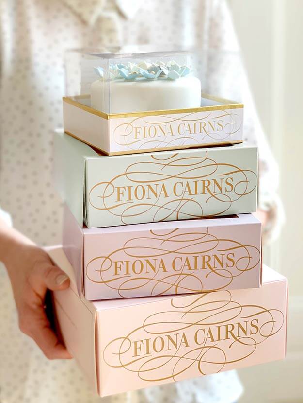 Tower of Fiona Cairns cake packaging. Pastel colours. Gold foil logo. Held in hands. Design by Andsome Ltd
